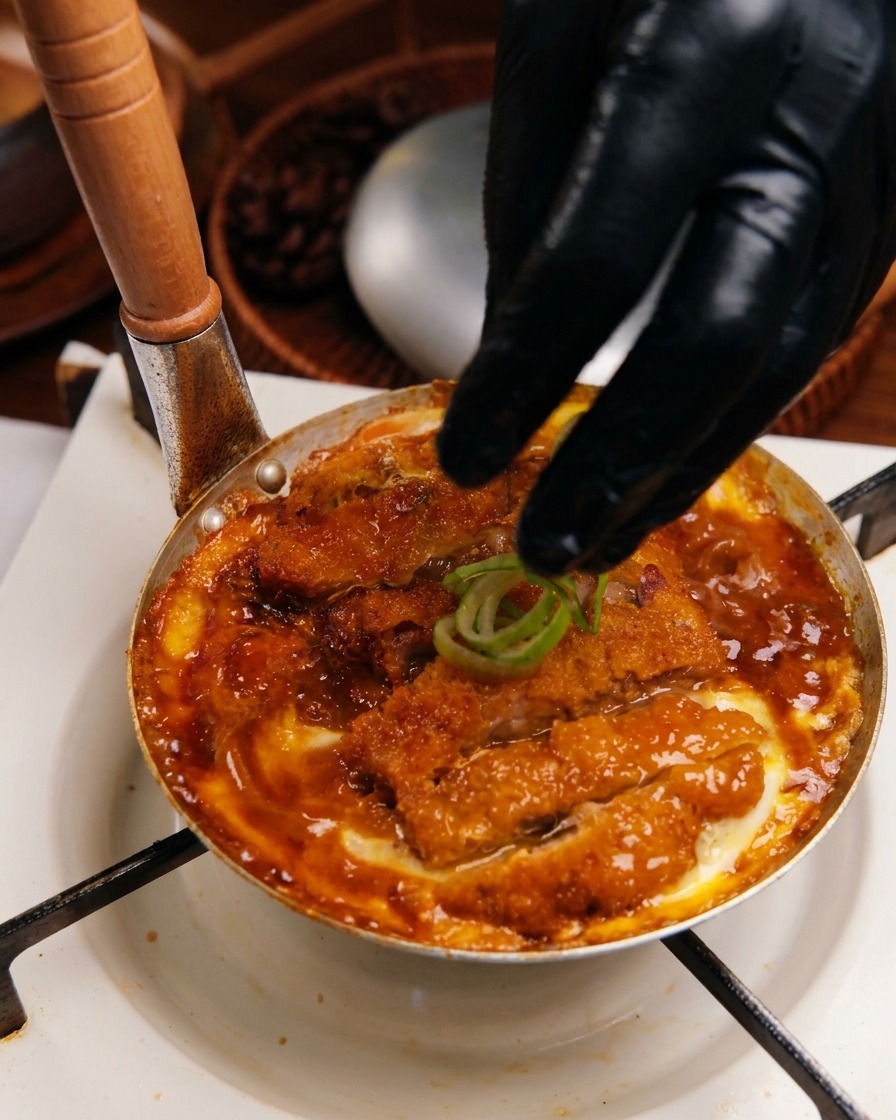 Main gantée déposant des rondelles doignons verts frais sur le mélange Katsudon cuit dans une poêle.