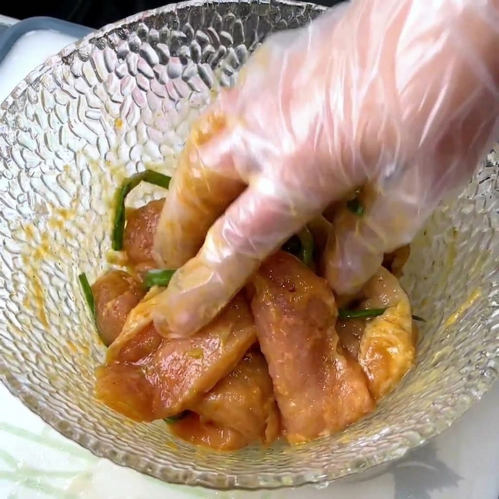 Une main gantée mélange les tranches de porc crues avec les cébettes et la marinade liquide dans un bol en verre texturé.