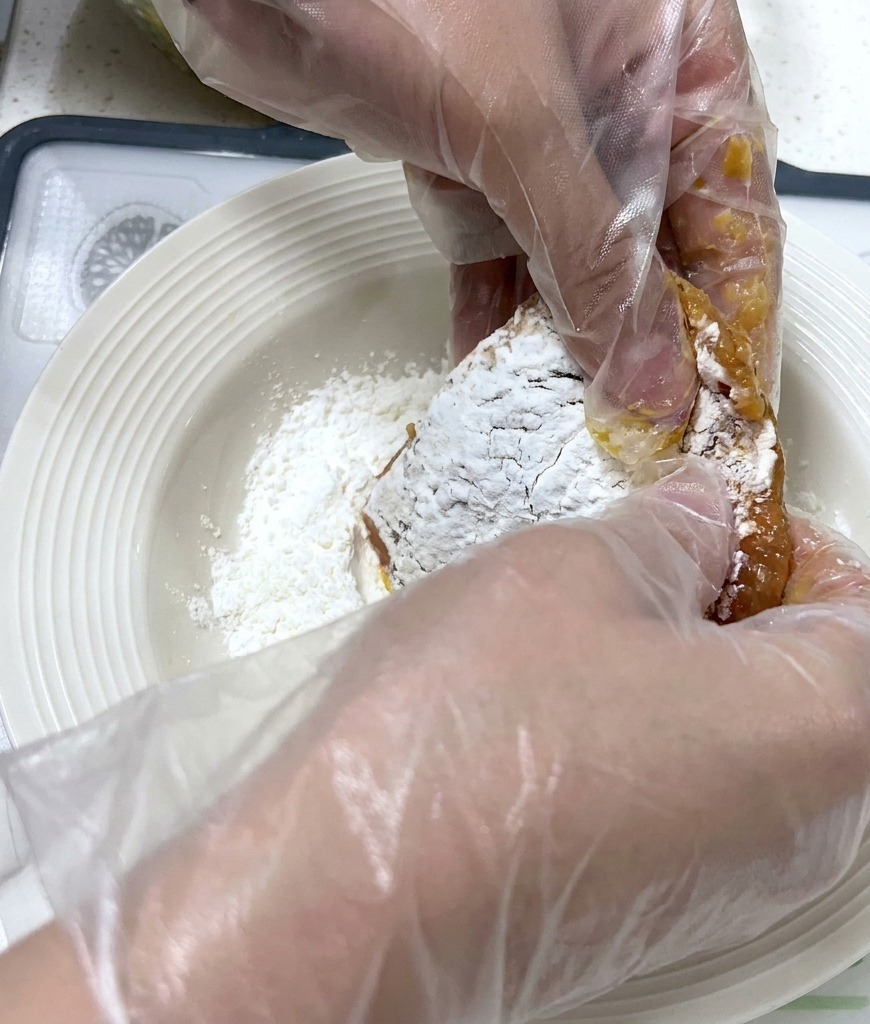 Des mains gantées pressent une tranche de porc marinée dans une assiette creuse remplie de fécule blanche sèche.