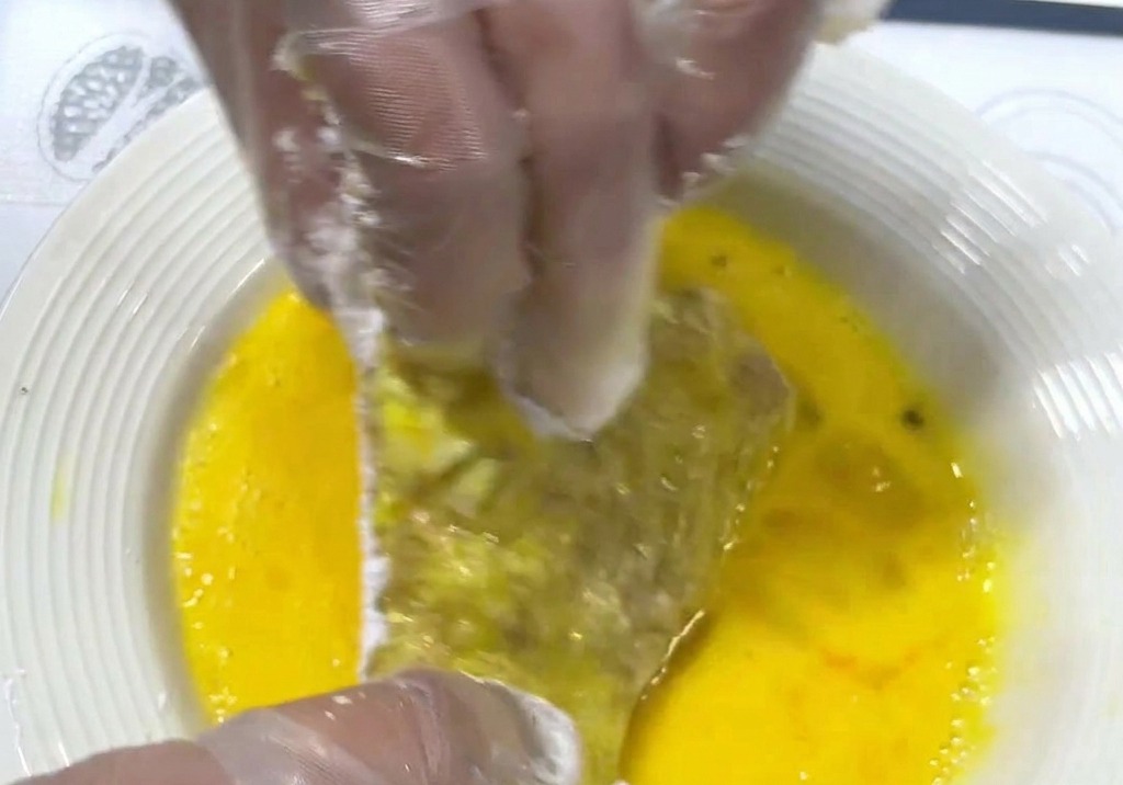 Des mains trempent une côtelette de porc farinée dans une assiette contenant un mélange dœufs battus dun jaune éclatant.