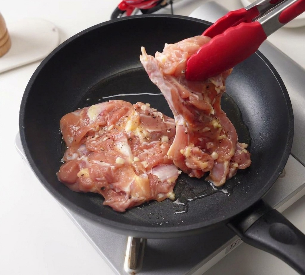 Une pince déposant un haut de cuisse de poulet mariné dans une poêle noire contenant un filet dhuile chaude.