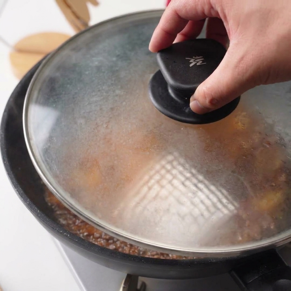 Une main posant un couvercle en verre sur une poêle contenant le poulet et la sauce qui mijote.