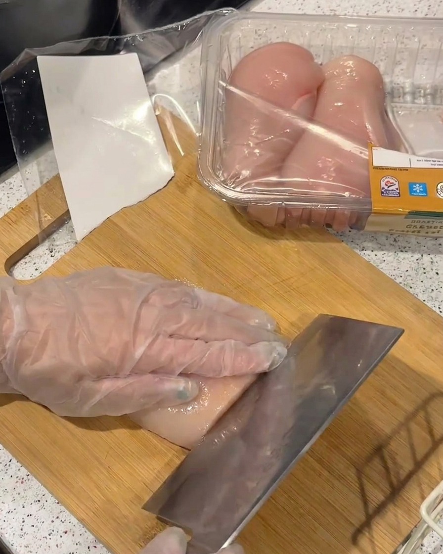 Une main gantée tranchant un blanc de poulet en filets fins avec un large couteau sur une planche en bambou.