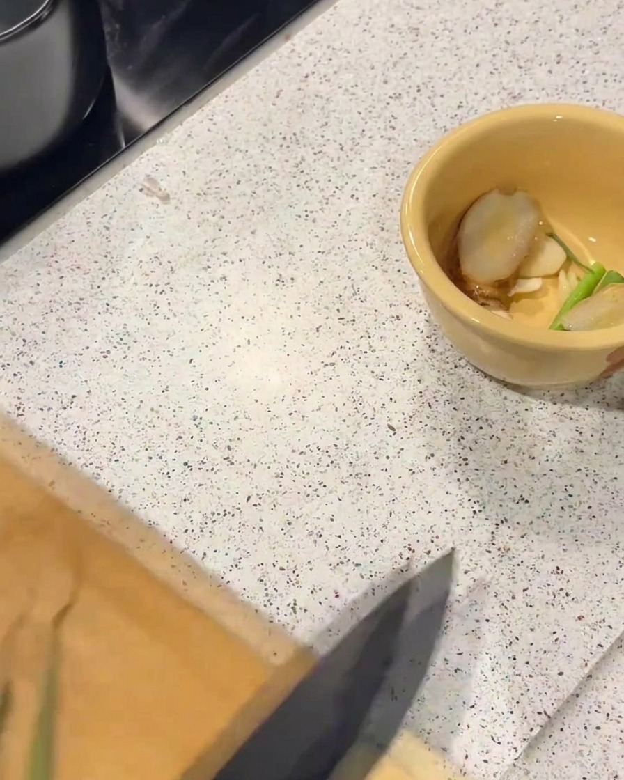 Un bol en céramique jaune contenant des tranches de gingembre et des morceaux doignons verts sur un plan de travail.