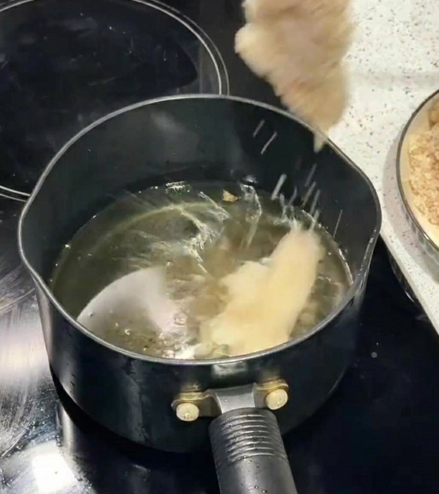 Une main utilisant des baguettes pour déposer un filet de poulet pané dans une huile de friture bouillonnante.