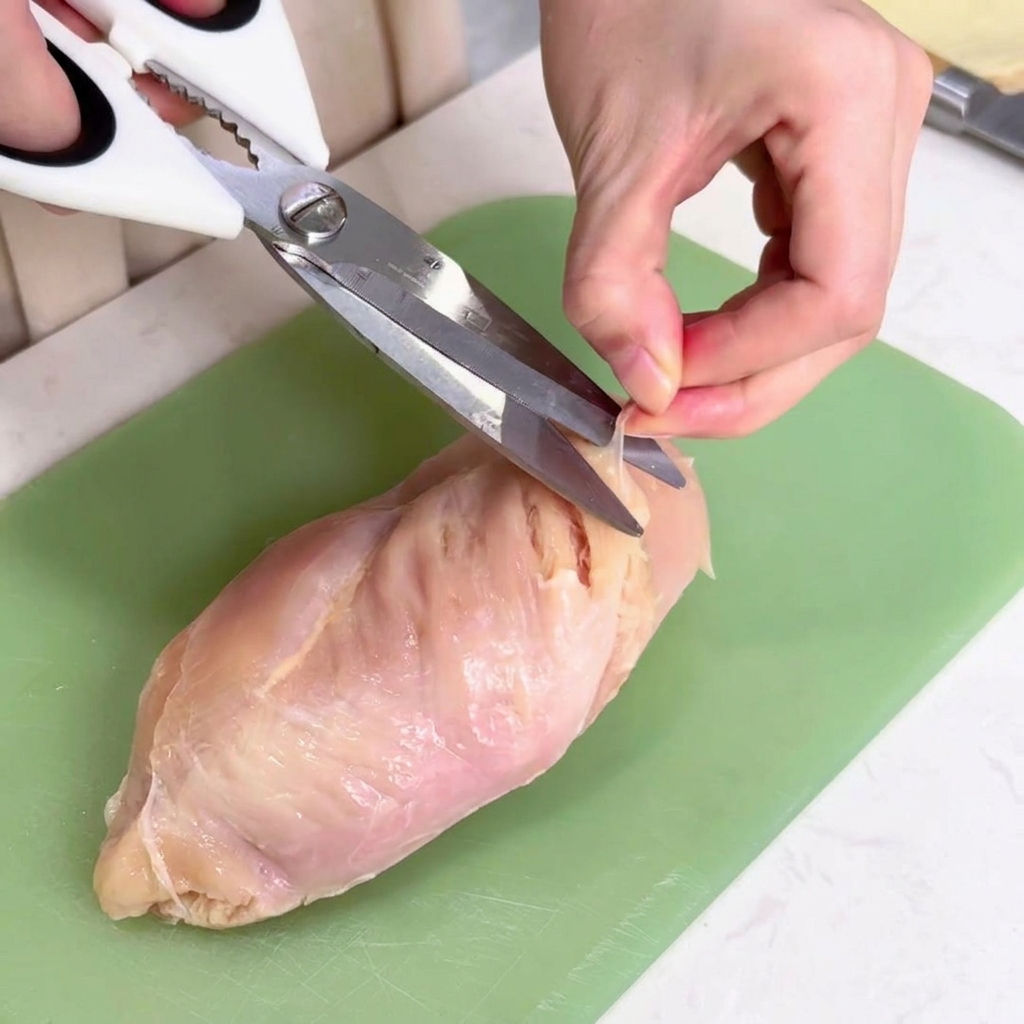 Une main utilisant des ciseaux de cuisine pour couper la fine membrane translucide dun blanc de poulet sur une planche verte.