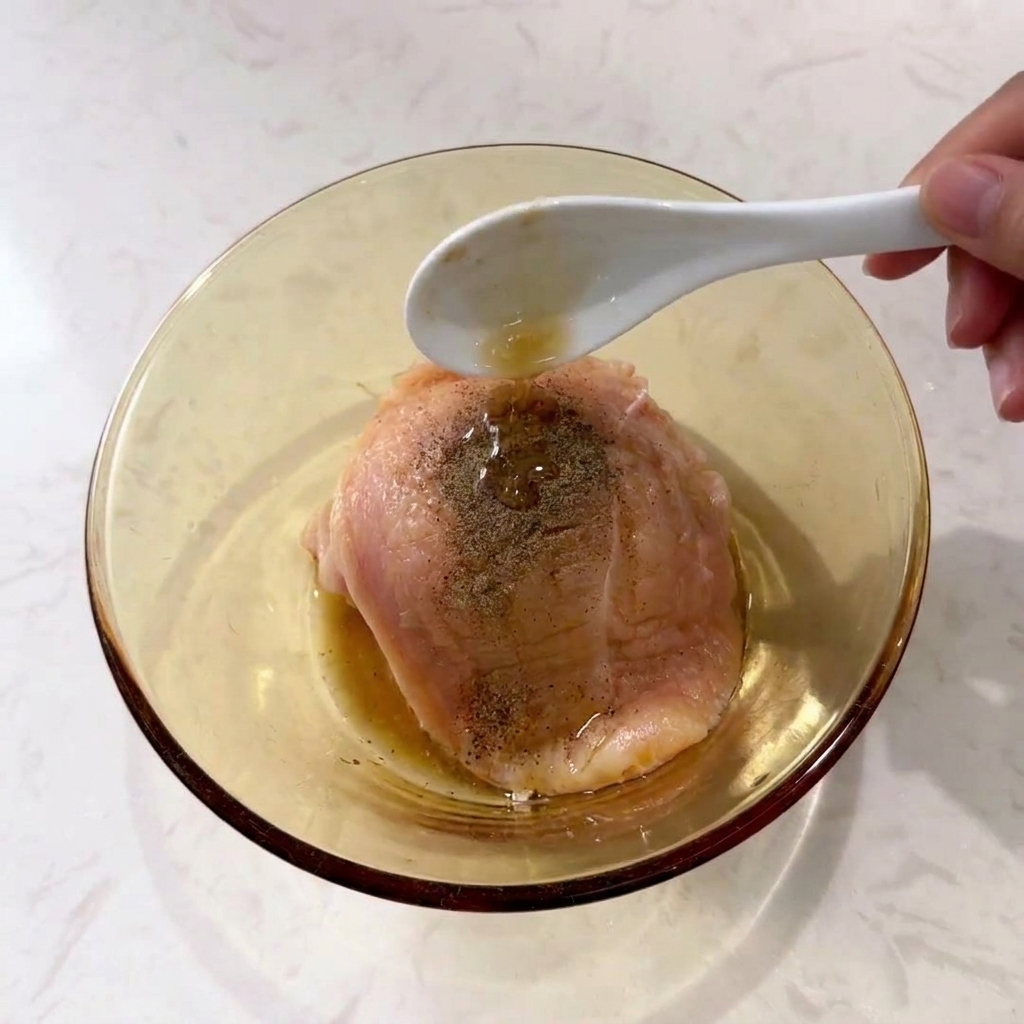 Une cuillère versant de lassaisonnement liquide sur un blanc de poulet saupoudré de poivre noir dans un bol en verre.