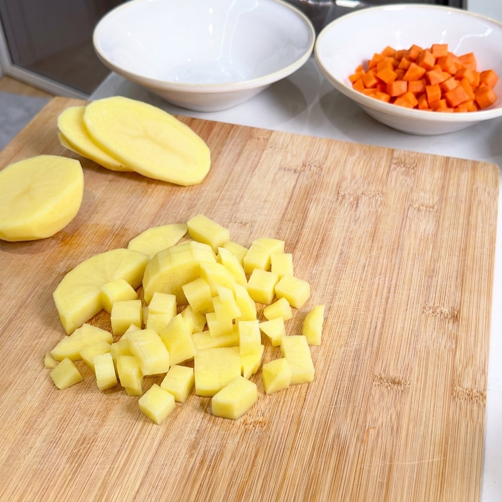 Cubes de pommes de terre jaunes empilés sur une planche à côté dun bol de carottes coupées.