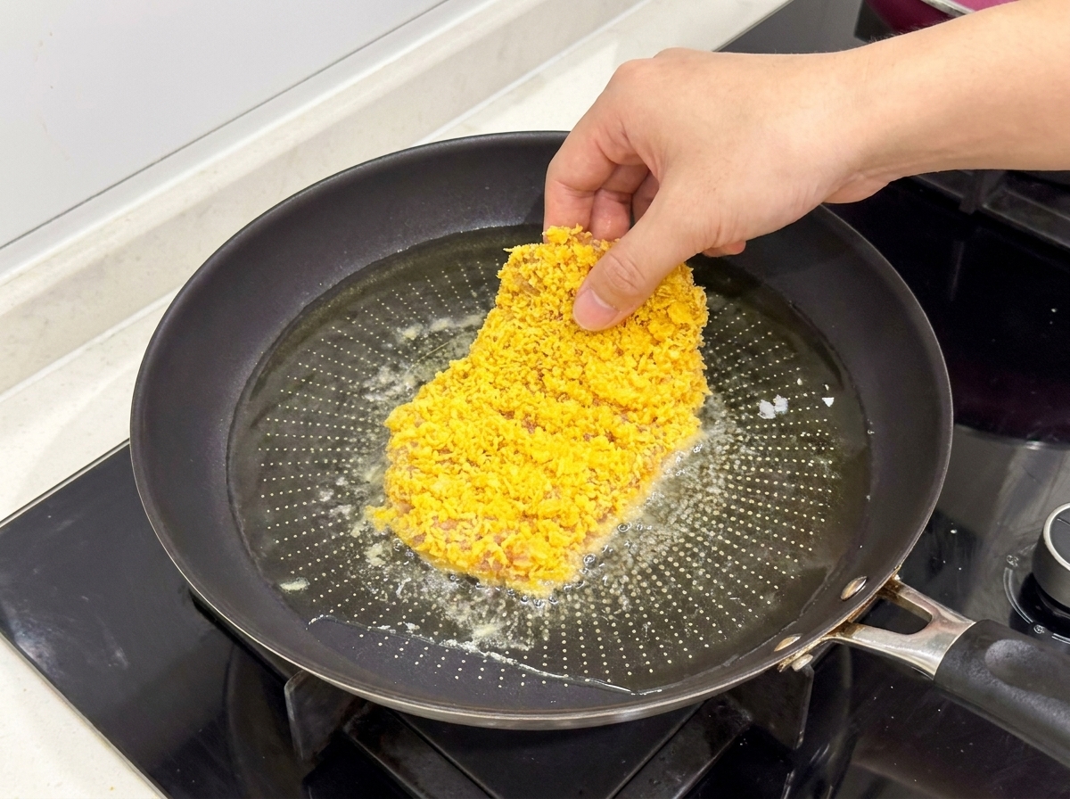 Une escalope de poulet panée déposée dans une poêle contenant de lhuile chaude et frémissante.