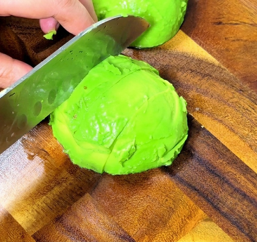 Gros plan sur des mains utilisant un couteau professionnel pour couper un avocat pelé en dés réguliers sur une planche en bois.