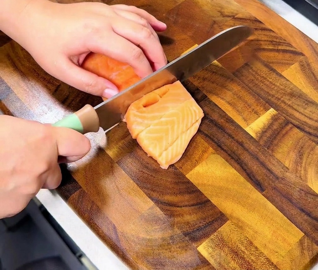 Mains tranchant un filet de saumon frais orangé sur une planche en bois pour créer des cubes uniformes.