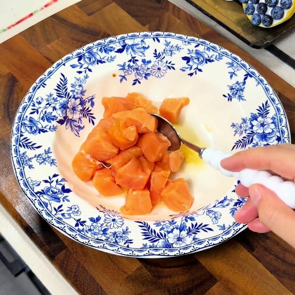 Une main mélangeant les cubes de saumon dans un bol bleu et blanc à laide dune cuillère blanche pour bien les enrober de marinade.
