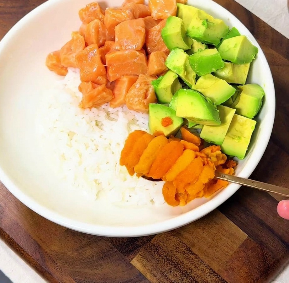 Une main déposant des morceaux doursin orange vif sur un bol de riz garni de saumon et davocat.