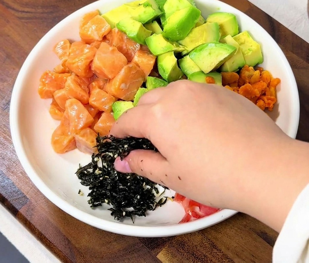 Une main saupoudrant des lanières dalgues nori séchées sur une partie vide du bol de riz à côté du saumon.