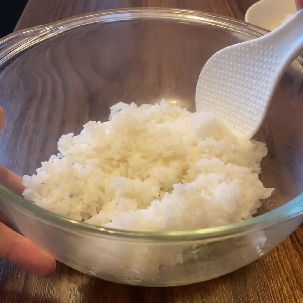 Une spatule à riz texturée mélangeant du riz blanc fumant dans un grand saladier en verre.