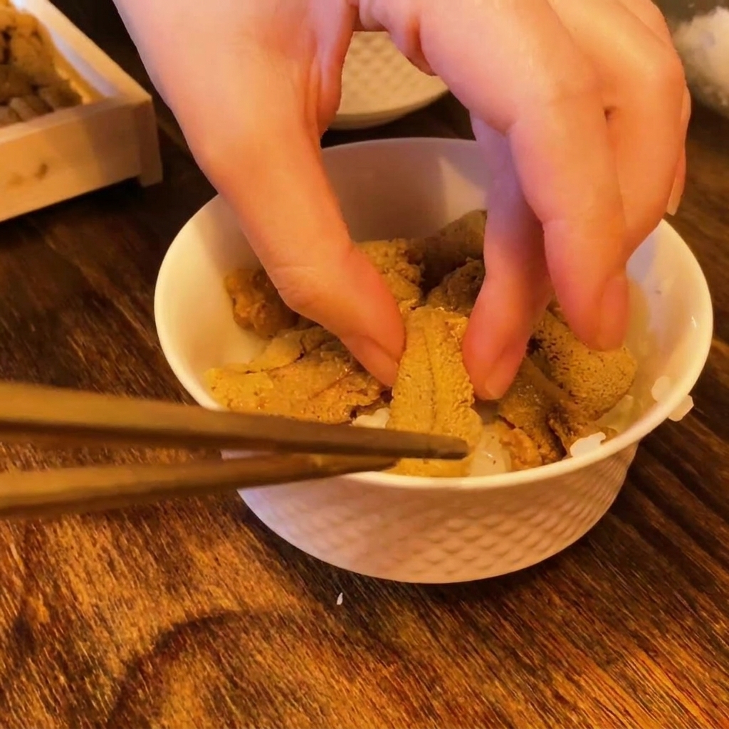 Des baguettes et des doigts déposant des morceaux doursin orange vif sur un lit de riz à sushi blanc.