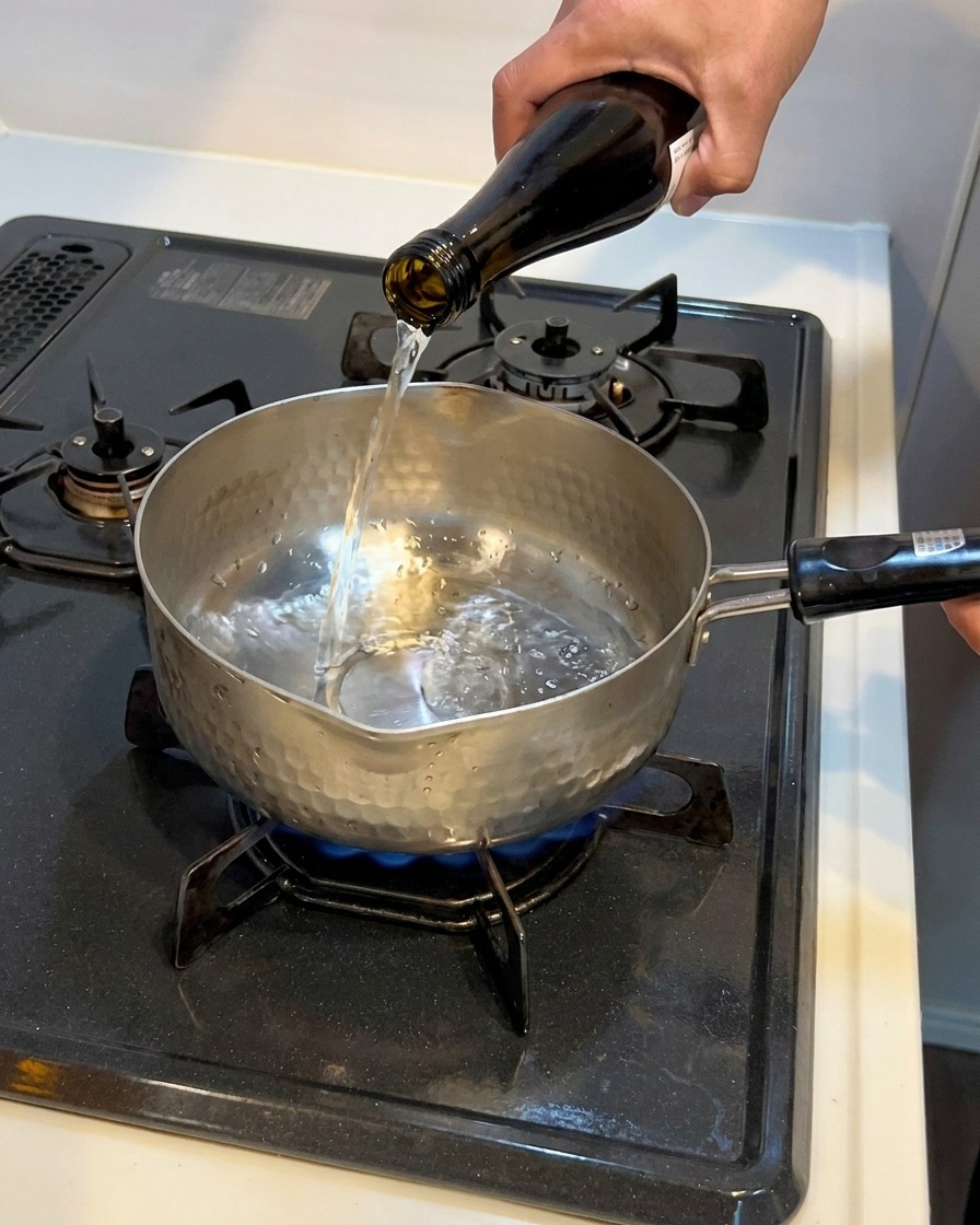 Liquide clair versé dune bouteille en verre sombre dans une petite casserole en métal sur une cuisinière à gaz.