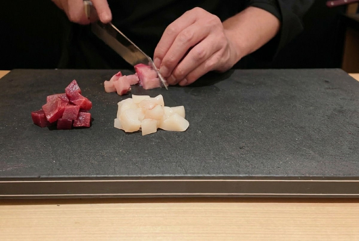 Un chef découpant du thon et de la sériole frais en petits dés réguliers sur une planche à découper noire.