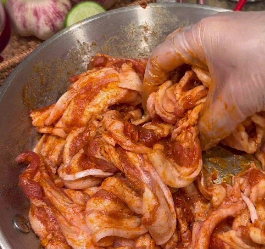 Une main gantée mélangeant des tranches de poitrine de porc avec une épaisse marinade rouge foncé.