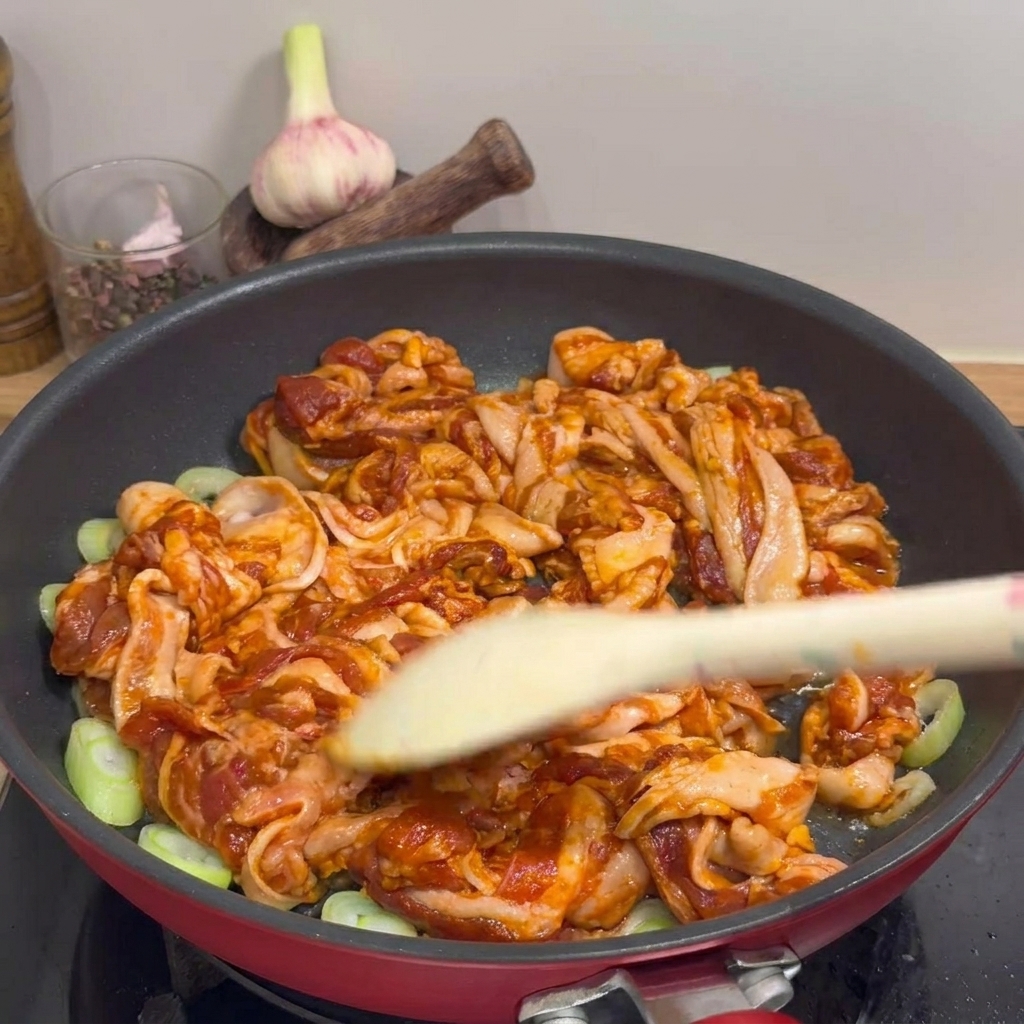 Plan rapproché de poitrine de porc sautée dans une poêle rouge avec des oignons verts, mélangée à laide dune spatule en bois.
