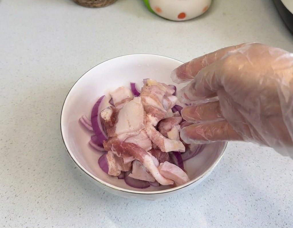 Une main portant un gant en plastique déposant des tranches de poitrine de porc crue dans un bol rempli de rondelles doignons.
