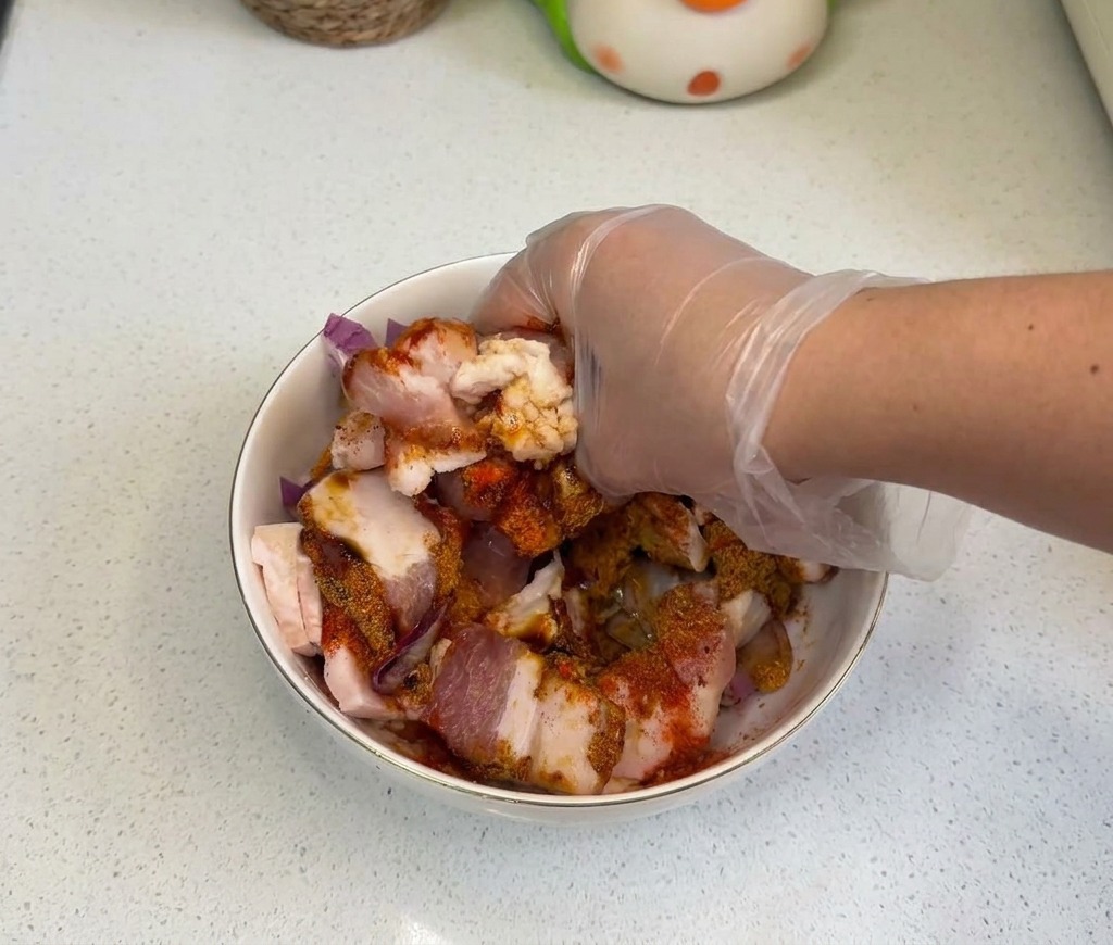 Une main gantée mélangeant vigoureusement la poitrine de porc, les oignons et les assaisonnements.