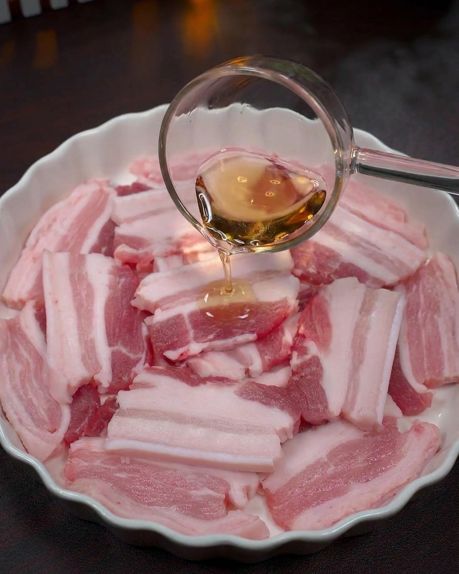 Liquide transparent versé depuis une louche en verre sur des tranches de porc crues dans un bol blanc.