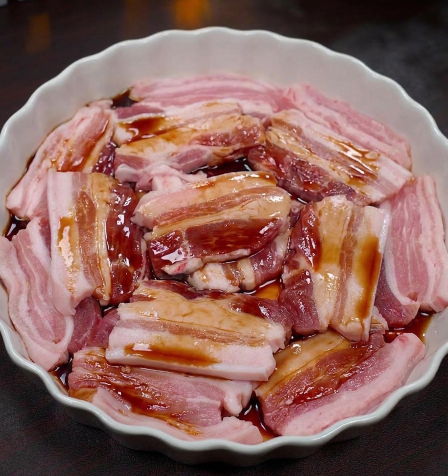 Tranches de poitrine de porc crue dans un plat blanc festonné recouvertes dune marinade foncée.