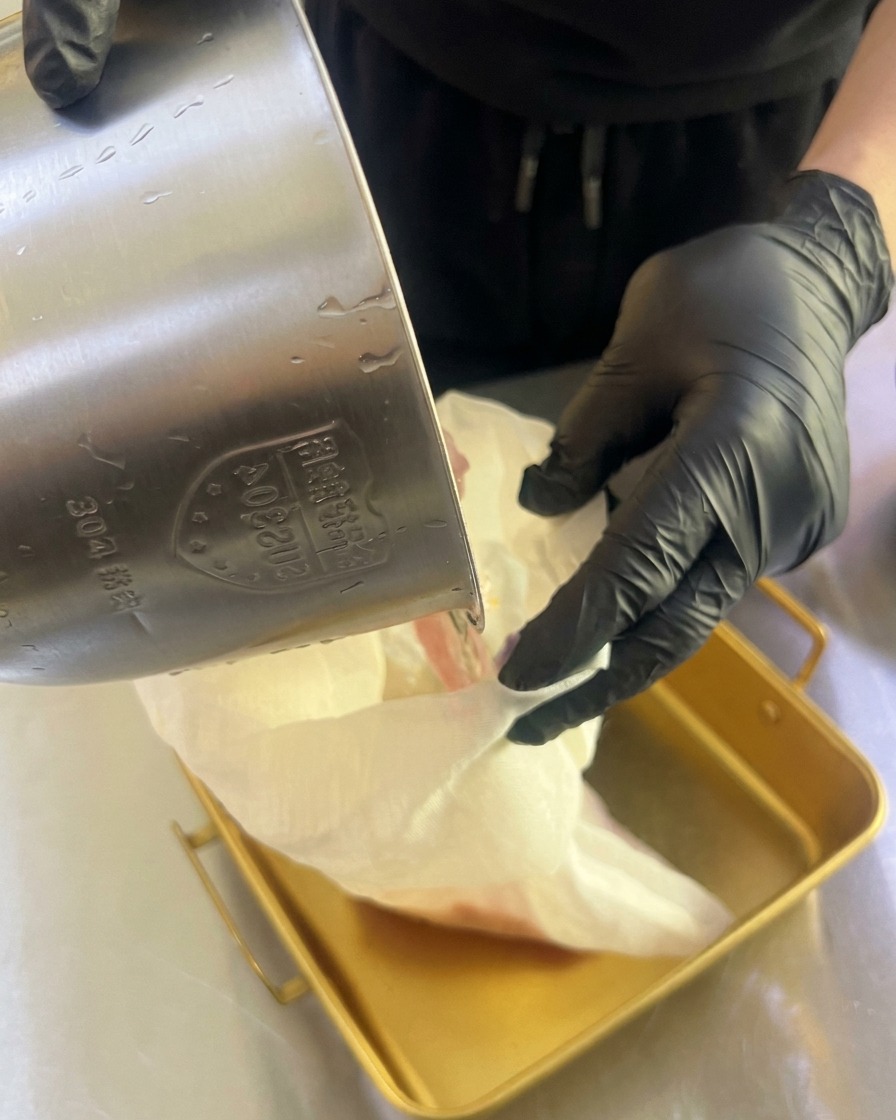Des mains gantées pressant un sac en étamine pour en extraire le jus de fruit frais dans un plateau métallique.