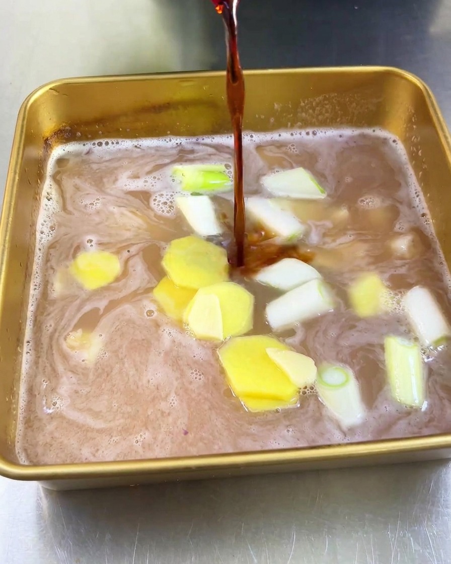 Un filet de sauce soja brune versé dans un plateau contenant le jus de fruit clair, le gingembre et les oignons verts.