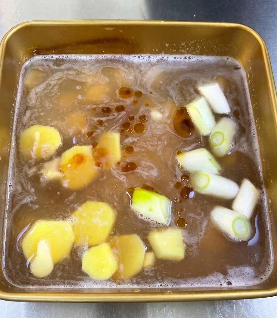 Vue de dessus dune marinade brune avec des gouttes dhuile de sésame flottant en surface parmi le gingembre et les cébettes.