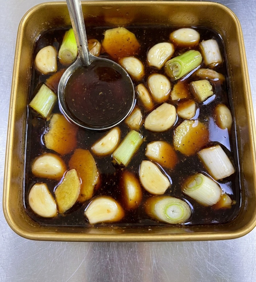Une louche en métal mélangeant une marinade sombre remplie de gingembre et doignons verts dans un plateau.