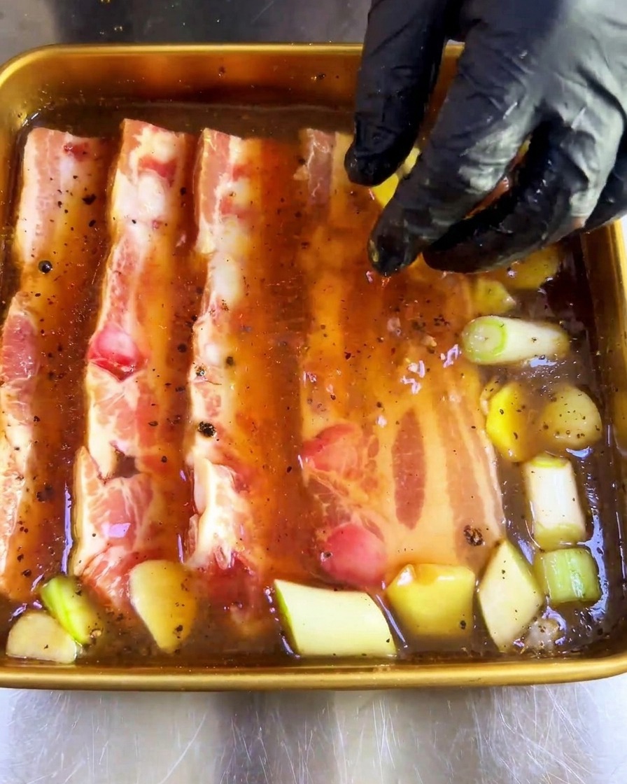 Une main gantée immergeant des tranches épaisses de porc cru dans un plateau de marinade brune.