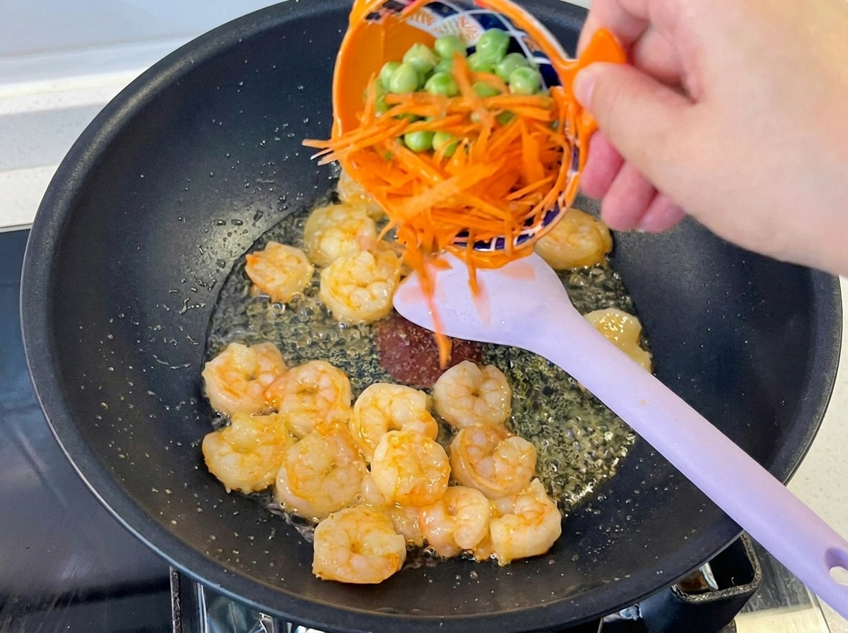 Une main versant des carottes râpées et des petits pois dun bol orange dans un wok contenant des crevettes et une spatule.