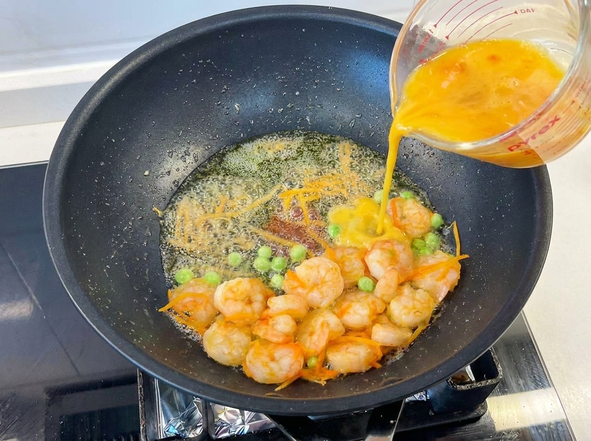 Œufs battus versés dun verre doseur dans une poêle chaude contenant crevettes, petits pois et carottes.