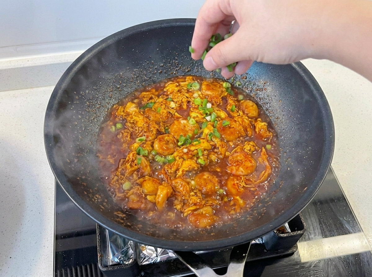 Une main parsemant des oignons verts hachés sur un plat de crevettes et œufs à la sauce vibrante dans une poêle.