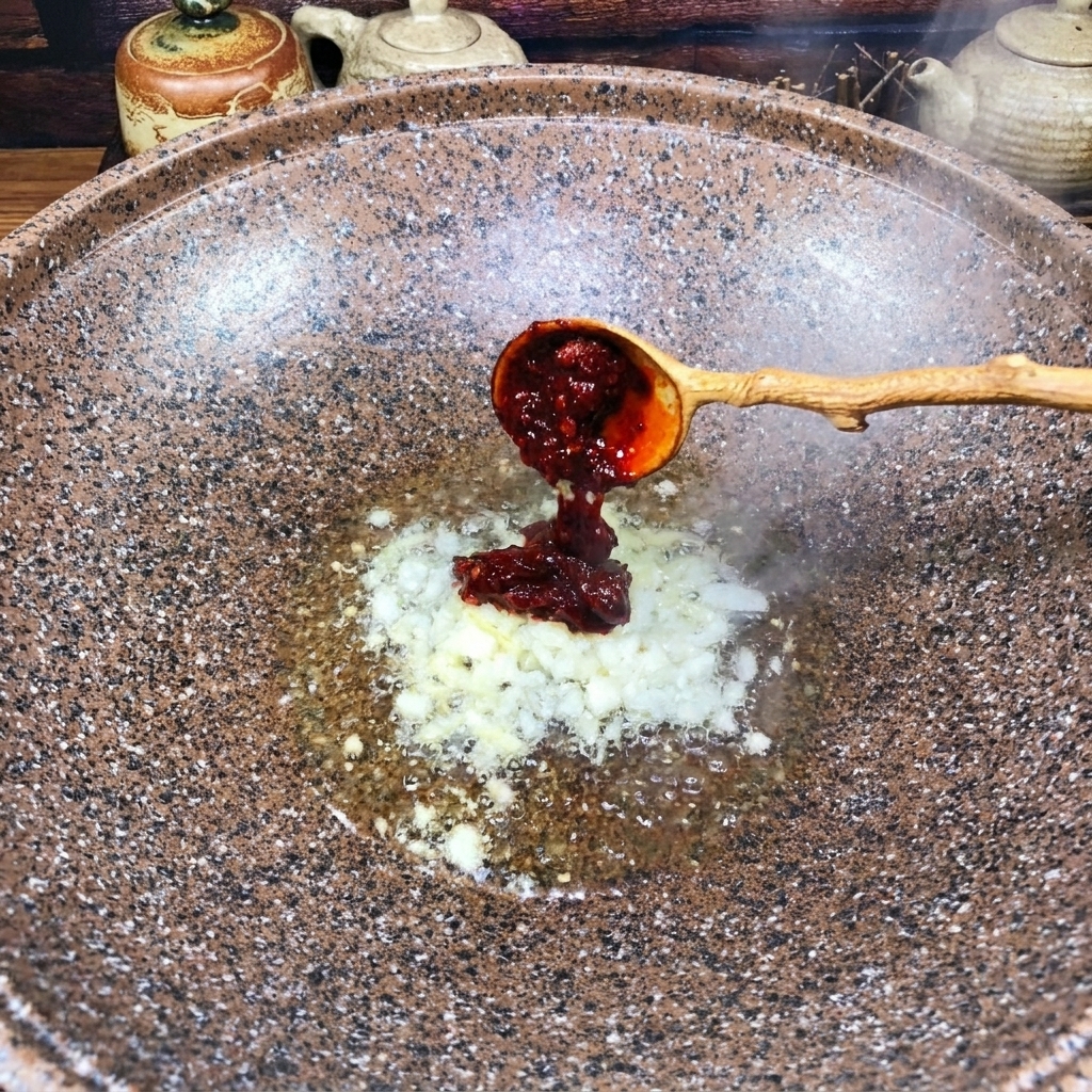 Une cuillère en bois ajoutant une dose de pâte de piment rouge foncé dans le wok avec les aromates.