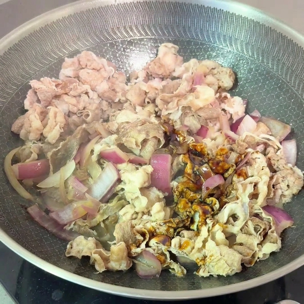 Tranches de bœuf blanchies et oignons mélangés à une sauce soja sombre dans une poêle.