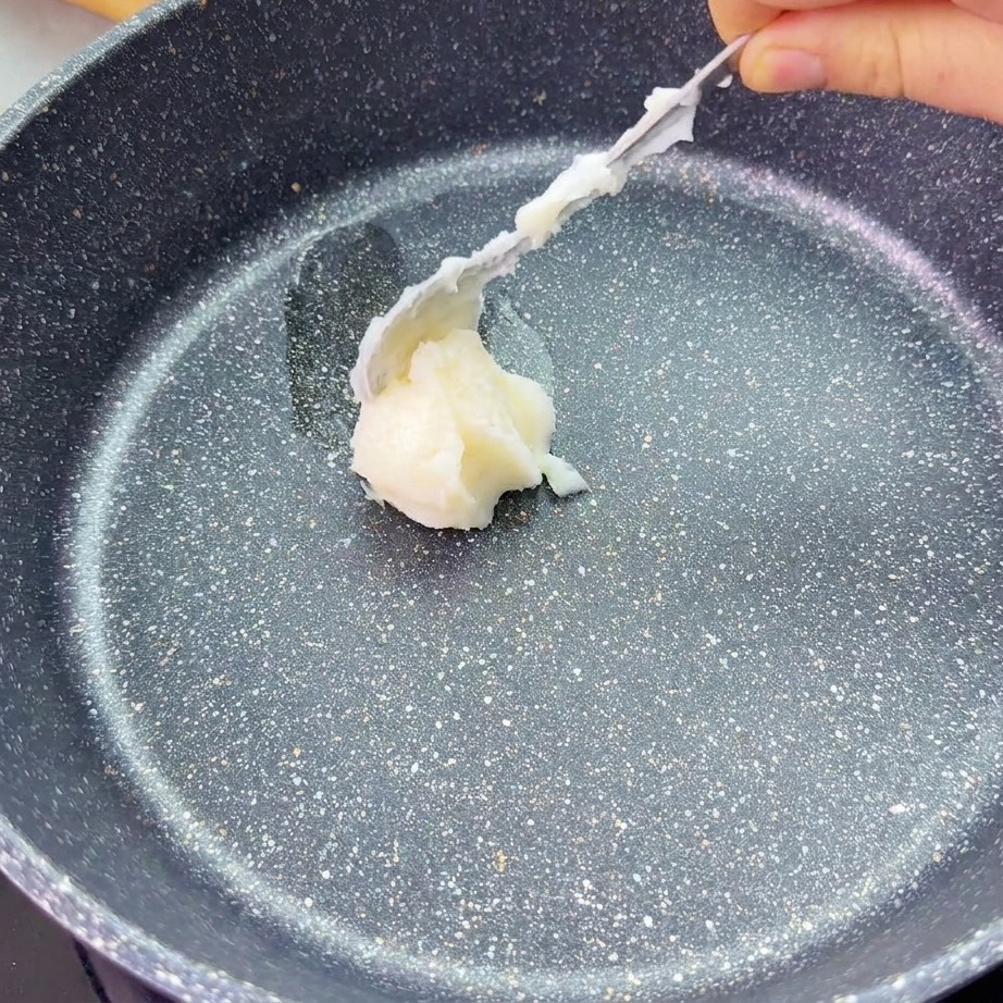 Une cuillère dépose une noisette de saindoux blanc au centre dune poêle sombre.