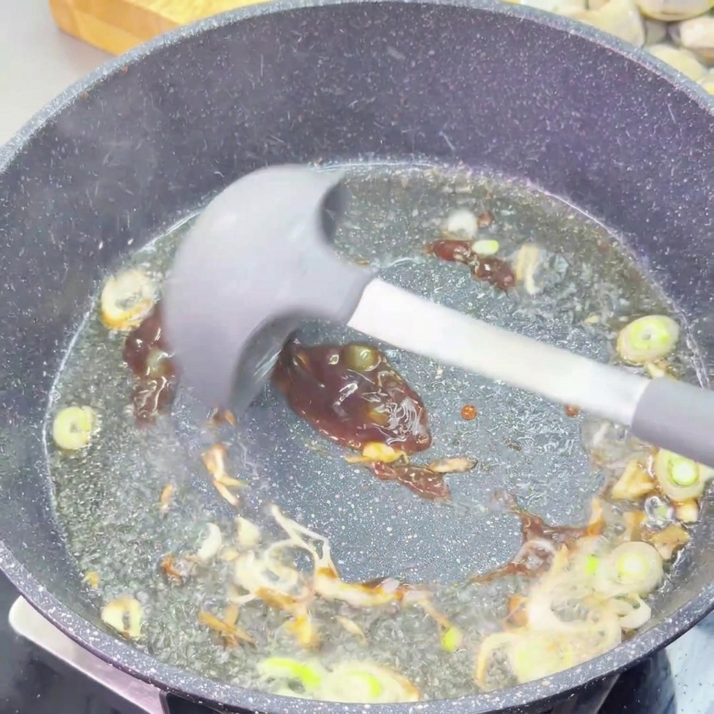 Une sauce dhuître épaisse et sombre est mélangée aux oignons verts dans une poêle avec une spatule en silicone grise.