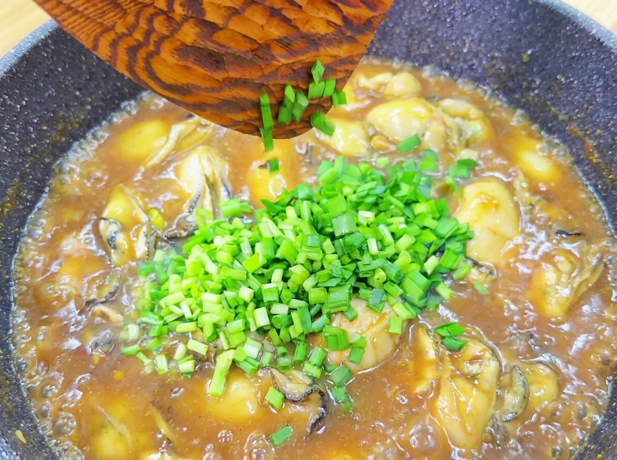 Ciboulette verte fraîchement hachée tombant dune spatule en bois dans une poêle dhuîtres en sauce.