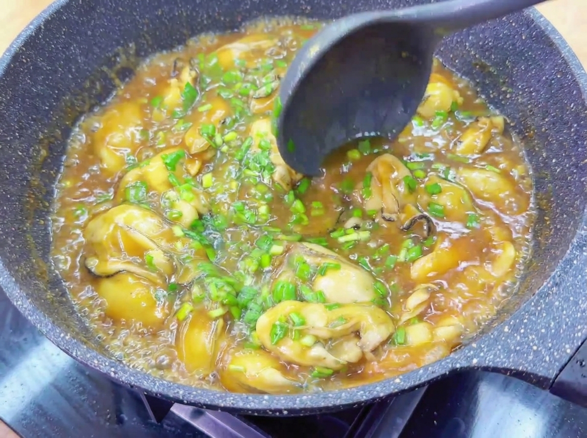 Une louche sombre mélangeant des huîtres entières et de la ciboulette dans une sauce brune brillante.
