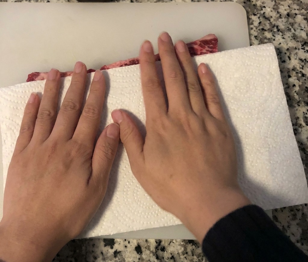 Mains utilisant de lessuie-tout pour absorber lhumidité de travers de bœuf crus posés sur une planche à découper blanche.
