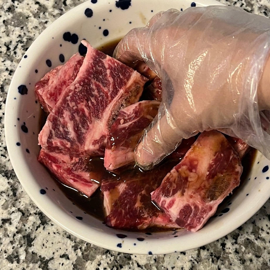 Une main gantée mélangeant les travers de bœuf avec la marinade sombre dans un bol pour assurer un enrobage homogène.