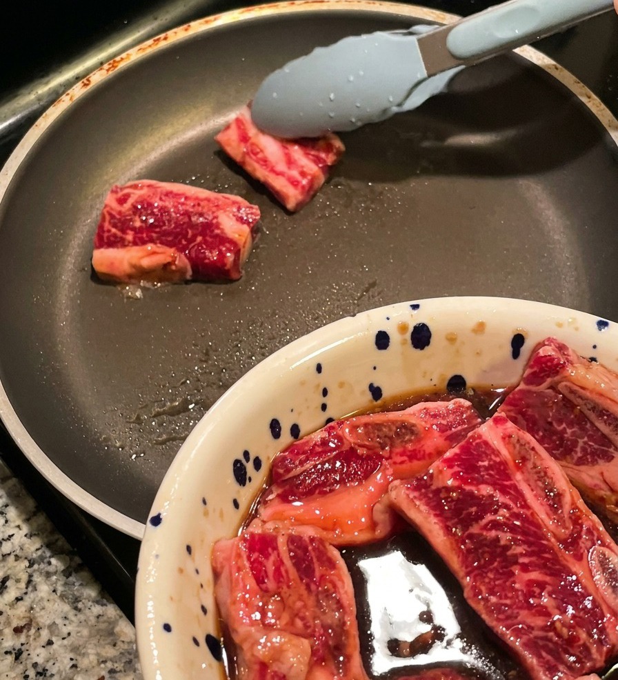 Pince déposant des tranches épaisses de travers de bœuf marinés dans une poêle chaude, avec le bol de viande au premier plan.