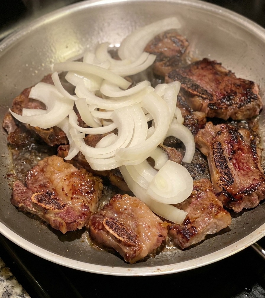 Un tas doignons blancs émincés posé sur les travers de bœuf dorés dans une poêle en inox.