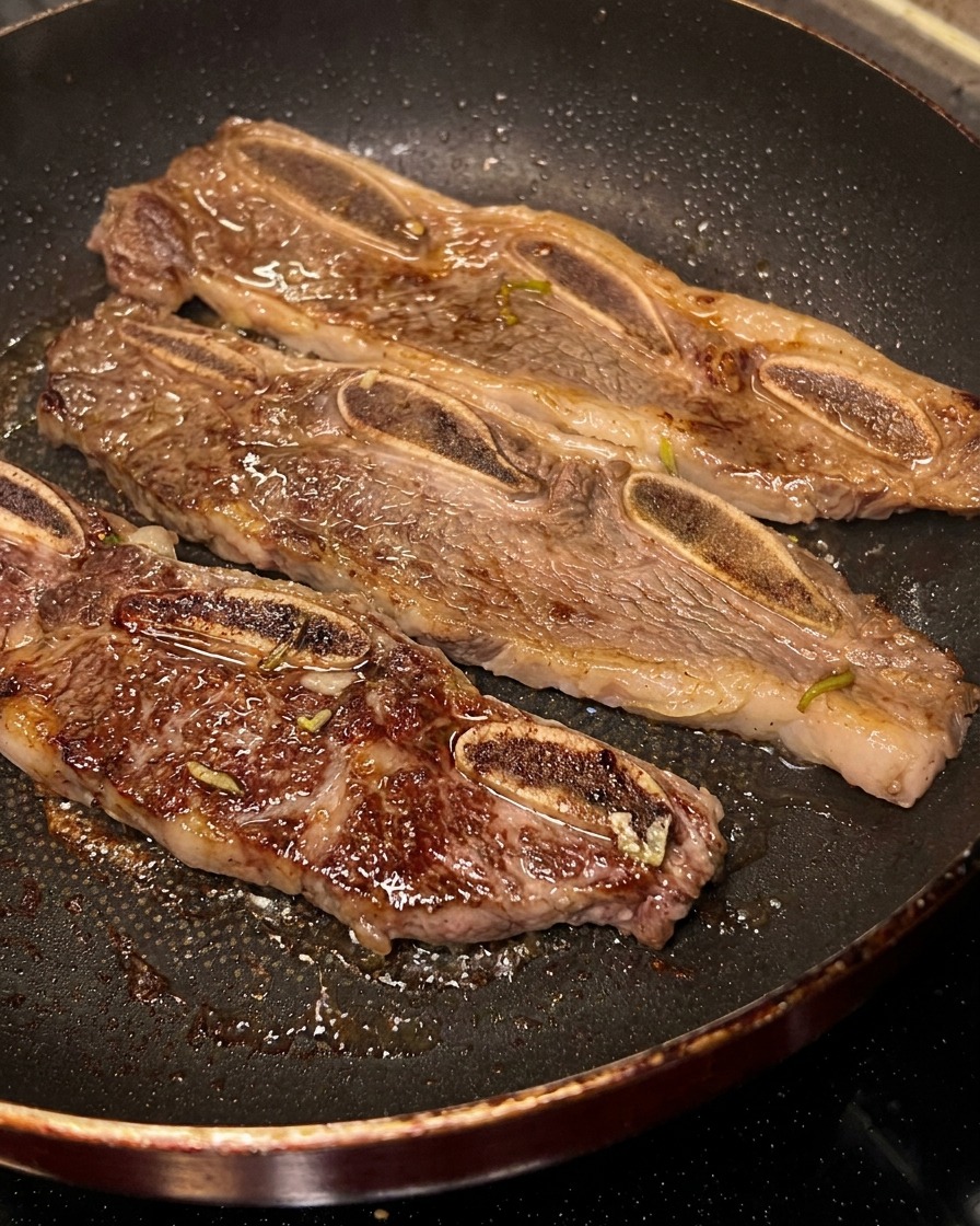 Trois tranches de LA Galbi grésillant dans une poêle, lune étant retournée pour montrer sa face caramélisée.