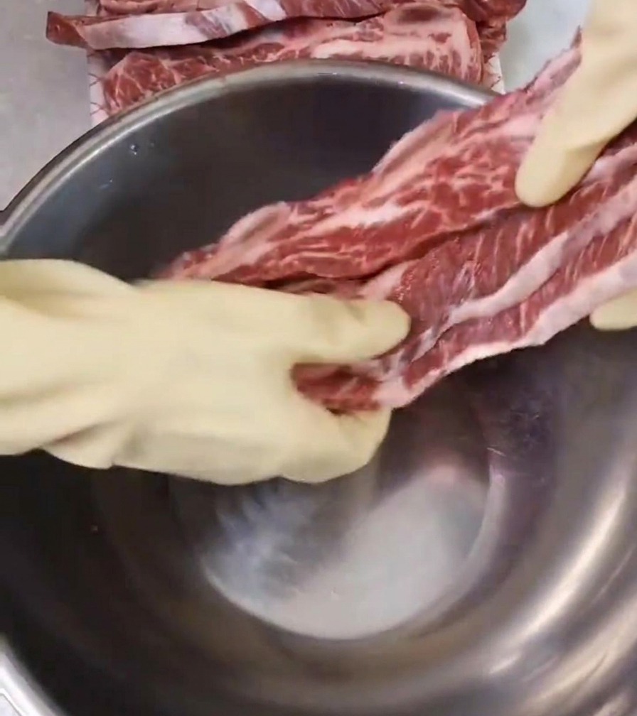 Une personne portant des gants blancs dispose des travers de bœuf marbrés dans un grand bol en inox.