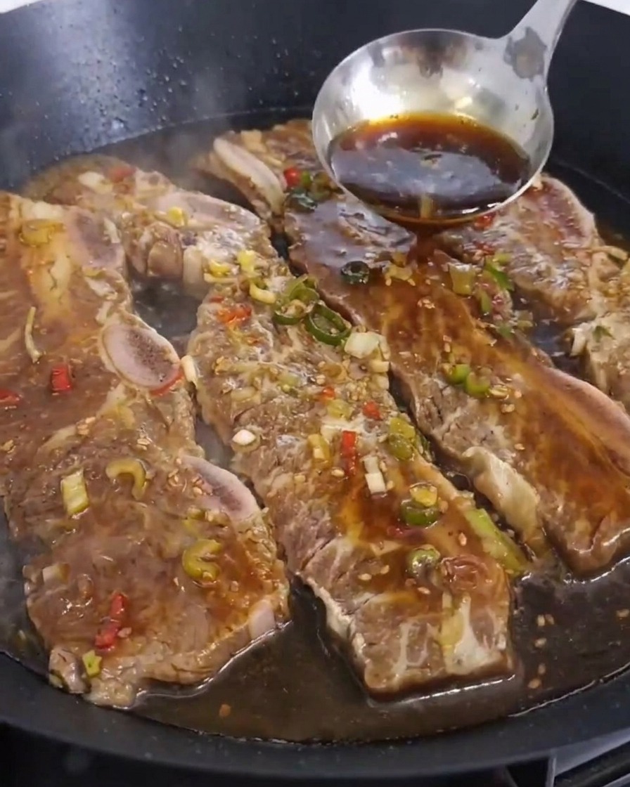 Une louche en inox verse une marinade sombre sur les travers de bœuf en train de griller.