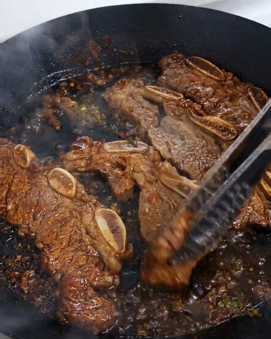 Travers de bœuf retournés avec des pinces dans une sauce sombre bouillonnante qui sépaissit.